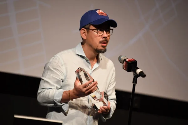 導演羅泓軫戴著藍色棒球帽與眼鏡，手拿紐約亞洲電影節的水晶獎座發言。