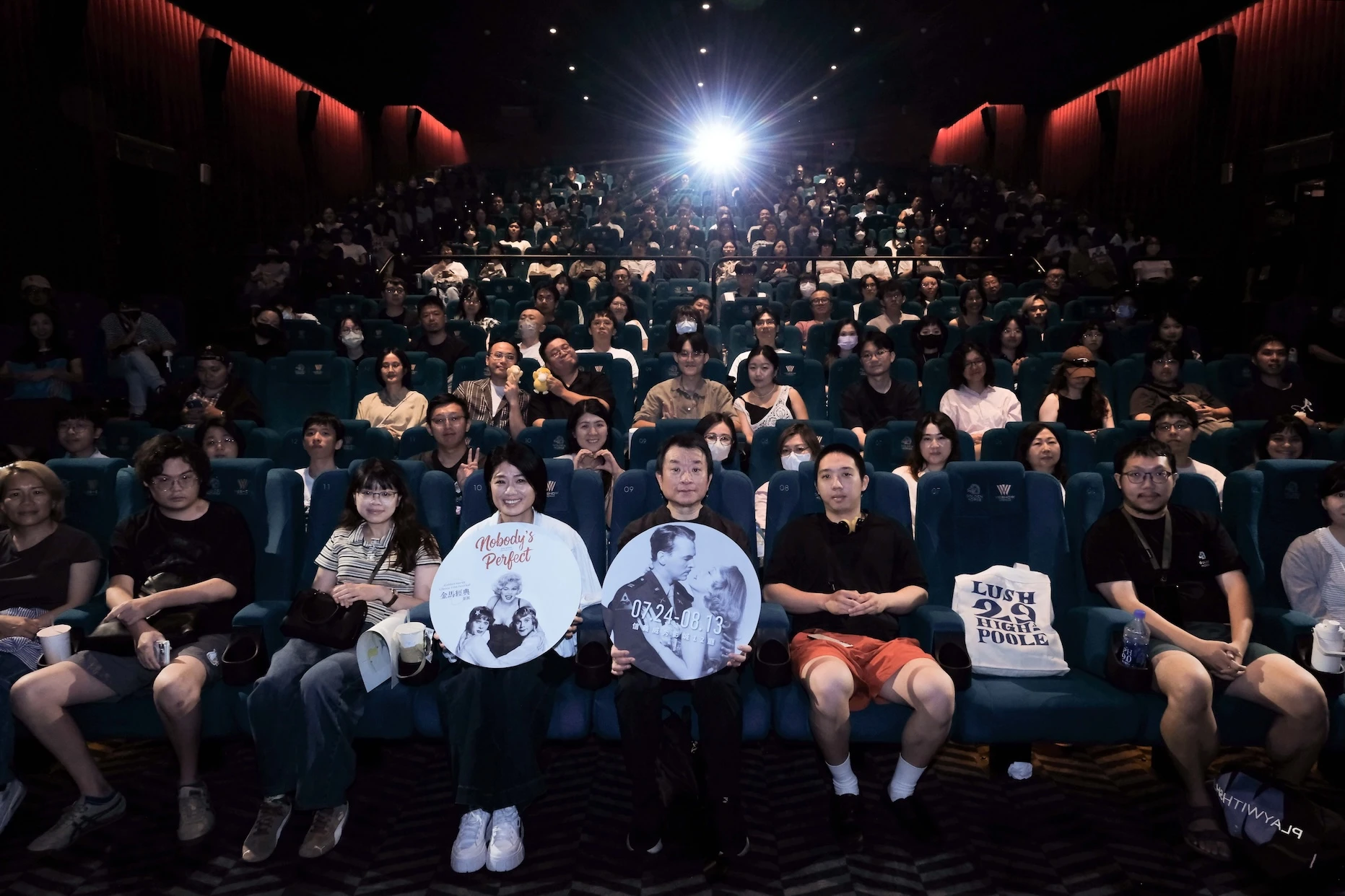 金馬影展執行長聞天祥與導演嚴藝文手持影展圓形宣傳牌，坐在影廳座位中央，與後方滿場的觀眾一同合影。