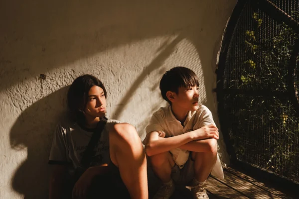 兩位少年坐在斑駁牆邊，陽光透過網狀結構灑落在他們身上。