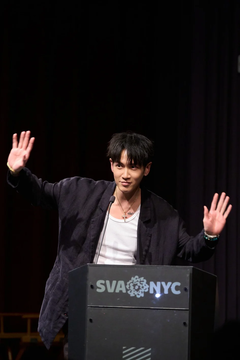 演員柯震東身穿深色西裝外套內搭白背心，站在講臺後方，雙手舉起揮手致意。