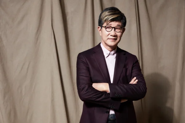 導演虞戡平身穿深色西裝與粉紅襯衫，雙手抱胸面對鏡頭微笑。
