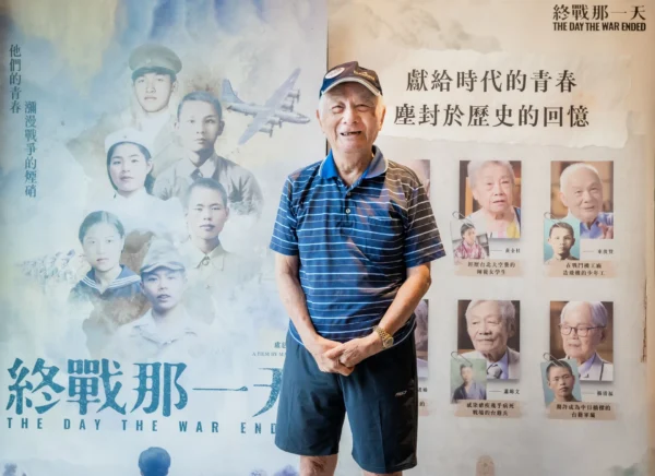 96歲受訪者東俊賢身穿藍白條紋POLO衫，站在《終戰那一天》青春版海報前微笑合影。