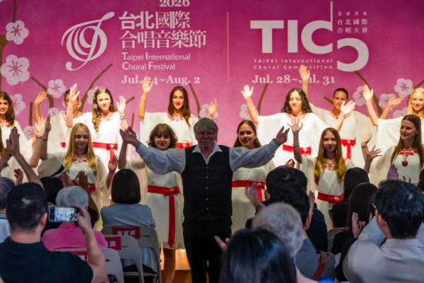 匈牙利 Cantemus 兒童合唱團成員身穿白色上衣與紅腰帶禮服，與指揮薩伯站在粉紅色背板前向台下致意，台下觀眾舉起手機拍照並鼓掌。
