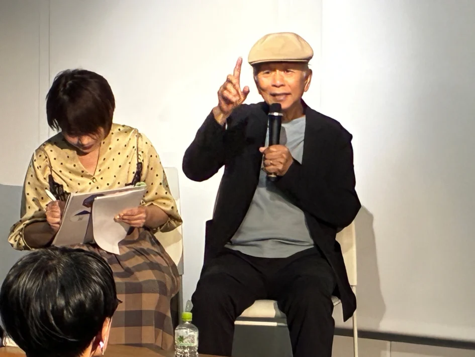攝影家阮義忠戴帽在台上拿麥克風發言，比出手指手勢