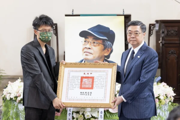 行政院長卓榮泰身穿西裝，右手持並遞出總統褒揚令給林榮基長子林靈均，林靈均戴著口罩與眼鏡、身穿深色西裝接過褒揚令，兩人前方擺放著褒揚令框架，背景為林榮基遺照。