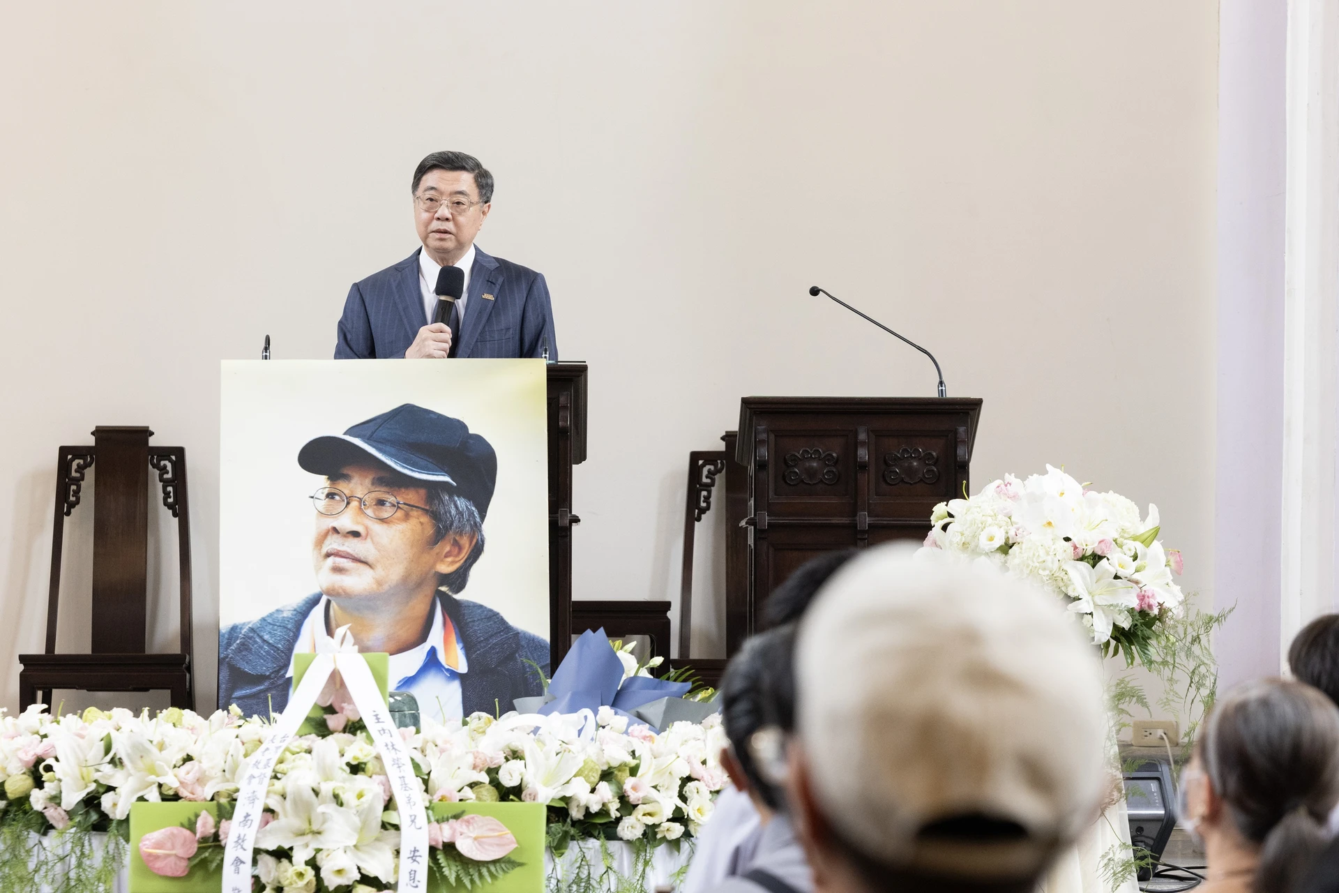 行政院長卓榮泰站在講臺前手持麥克風發言,講臺下方擺放著林榮基的遺照與白色花籃裝飾。