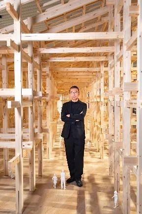 建築師藤本壯介穿著深色西裝站在木構造建築模型內部正中央,雙手交叉抱胸。