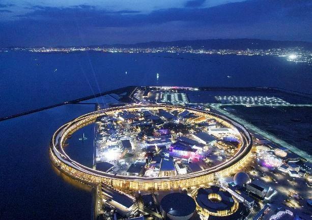 夜間空拍大阪・關西萬博會場,巨大的環形木造建築發出明亮燈光,環繞著展區與水岸。