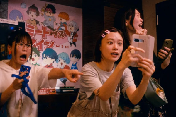 電影劇照中，女演員杉咲花與兩名女性友人拿著手機注視前方，露出驚訝表情，背景牆上貼有動漫角色海報。