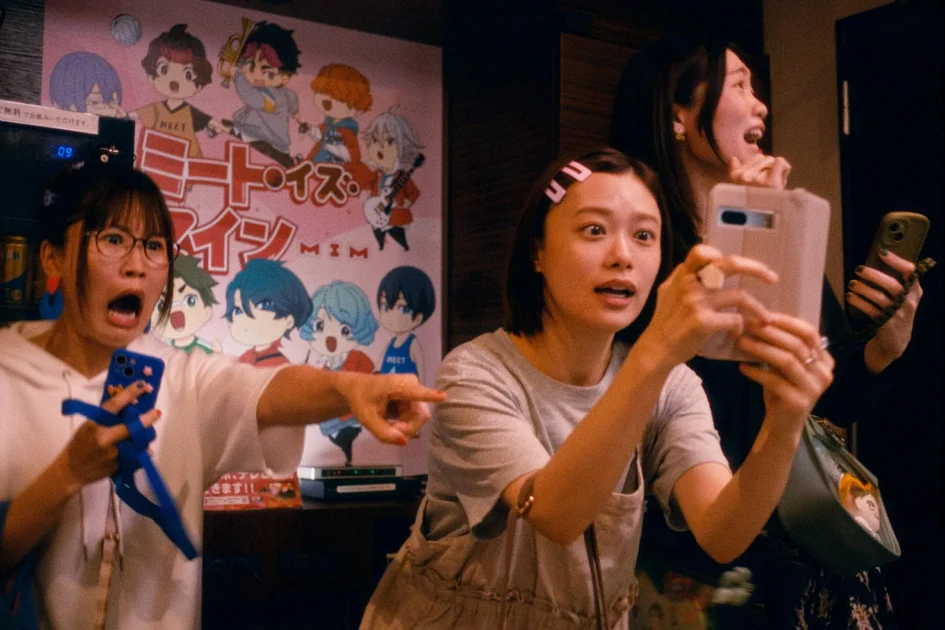 電影劇照中，女演員杉咲花與兩名女性友人拿著手機注視前方，露出驚訝表情，背景牆上貼有動漫角色海報。