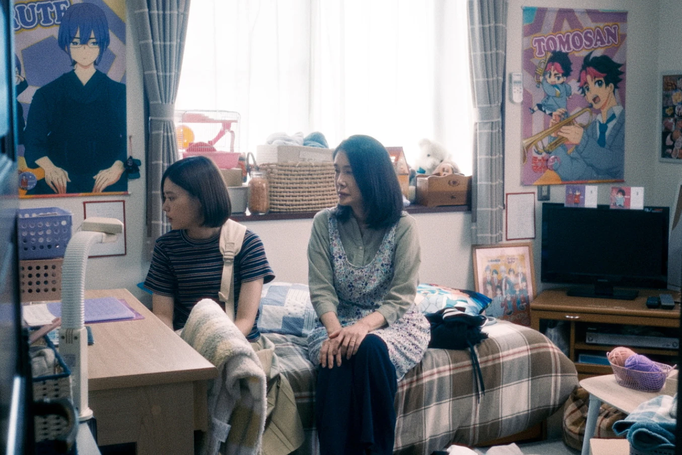電影劇照中,杉咲花坐在房間書桌旁,身旁坐著資深女演員,背景充滿動漫周邊與海報。