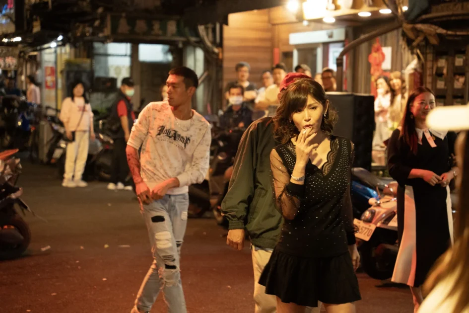 女演員鄭仲茵身穿黑色細肩帶上衣與短裙在街頭抽菸，背景可見多位路人與街景。