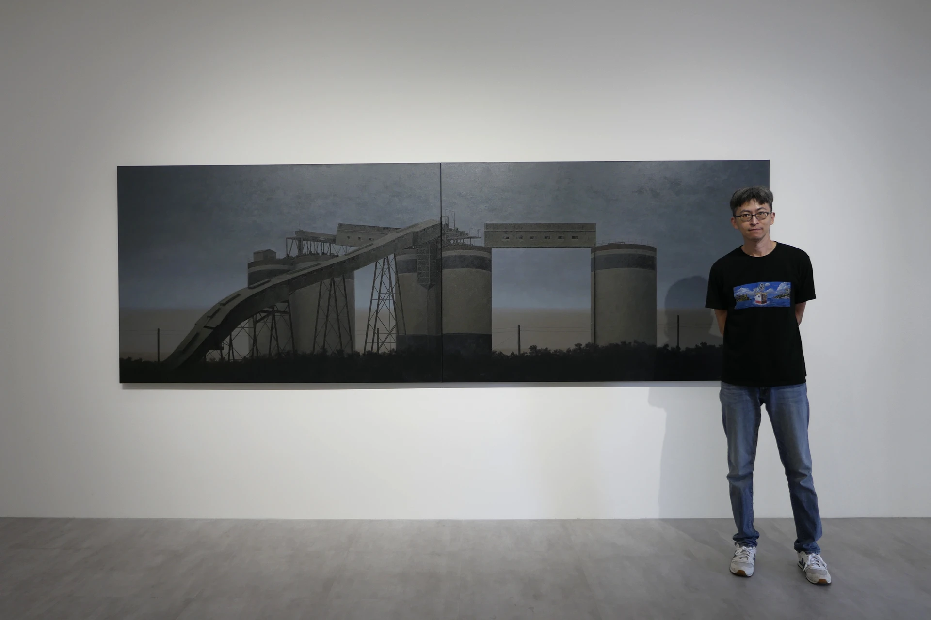 藝術家蔡孟閶站在大型油畫作品旁,身穿黑色T恤與牛仔褲,右手輕觸身側。