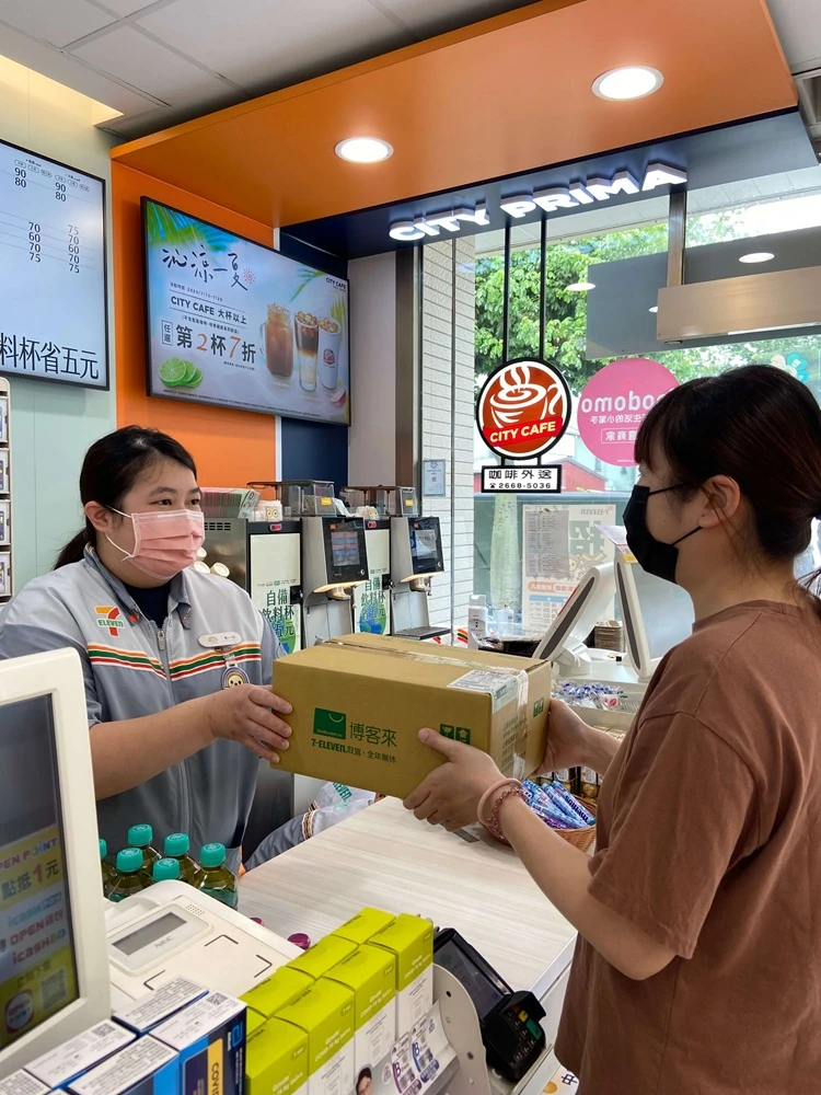 7-ELEVEN店員戴著粉紅色口罩，將博客來宅配紙箱遞給戴黑口罩的女性顧客。