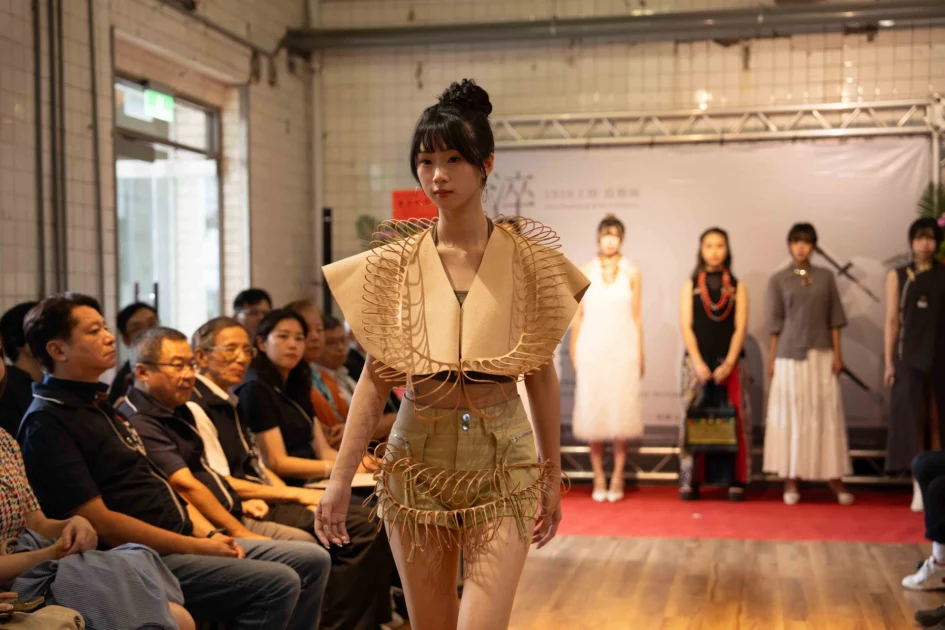 一名模特兒穿著具備獨特編織造型的服飾走在展場伸展台上，背景可見觀眾席與展覽背板。