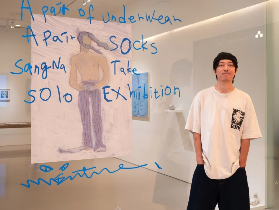 台灣潮流藝術家桑拿大可在展場中，右手插口袋站在印有展覽英文名稱的玻璃牆與畫作旁。