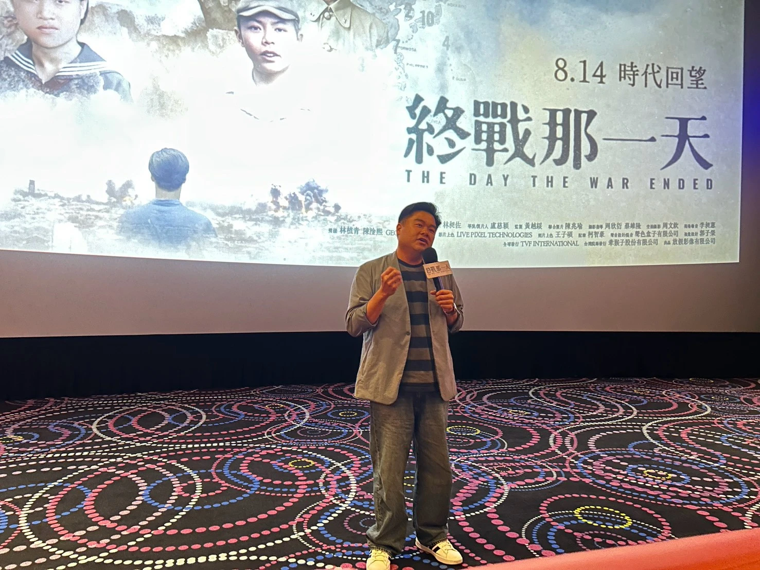 知名講師呂捷站在電影院銀幕前拿著麥克風發言,背景為電影宣傳海報。