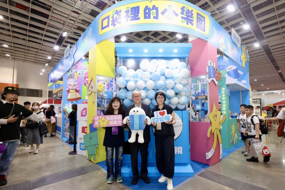 三位民眾與文化部長李遠站在藍色與粉色系的「口袋裡的小樂園」展位前，手拿宣傳牌與吉祥物玩偶合影，後方為大型扭蛋機造型裝置。