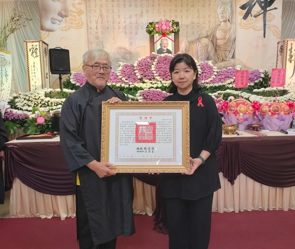 文化部政務次長李靜慧（右）與蕭在淦長子蕭維馨（左）手持總統褒揚令合影，兩人皆身穿黑色服裝，背景為告別式場地與蕭在淦遺照。