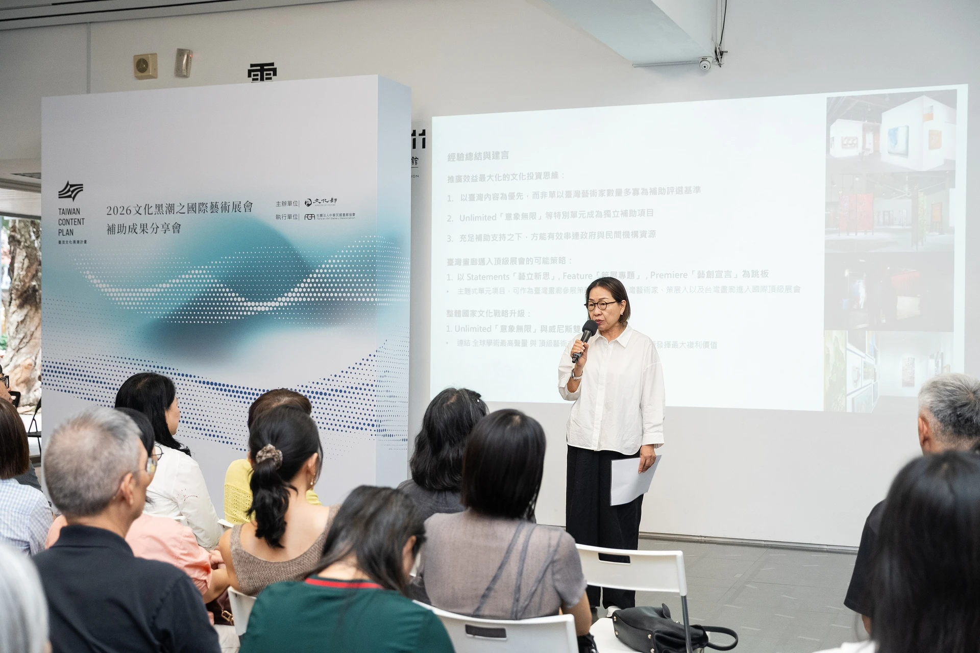 耿畫廊創辦人耿桂英手持麥克風於台前發表參展經驗,背後投影幕展示簡報資料。
