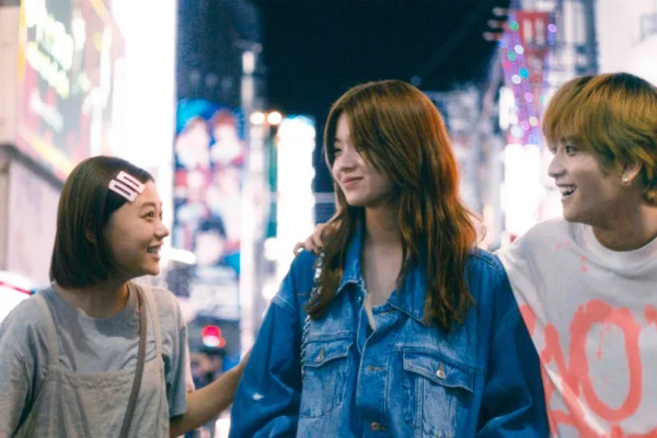 三名年輕男女站在夜晚亮著霓虹燈的街頭，左起為戴著髮夾的杉咲花、中間為長髮的南琴奈、右為金髮的板垣李光人，三人正微笑看著彼此。