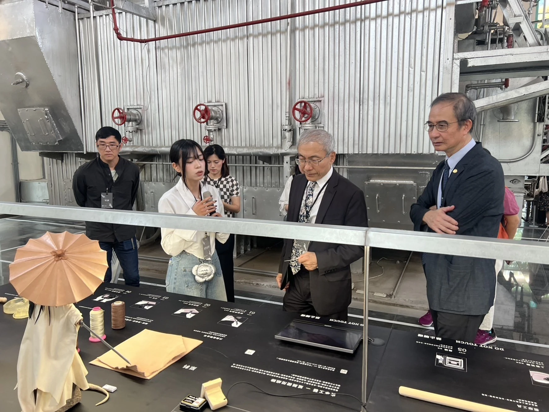 策展人員站在展位前向文化部文化資產局局長陳濟民與嶺東科技大學校長等貴賓介紹桌上的工藝創作素材與工具。