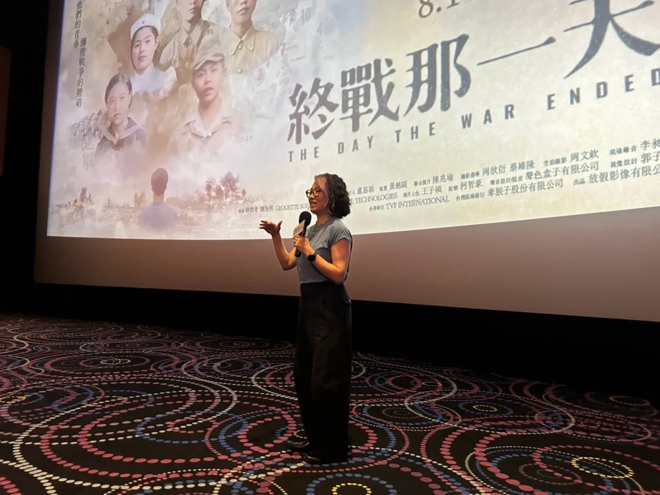 導演盧慈穎站在電影院銀幕前拿著麥克風講話，身後投影著電影海報。