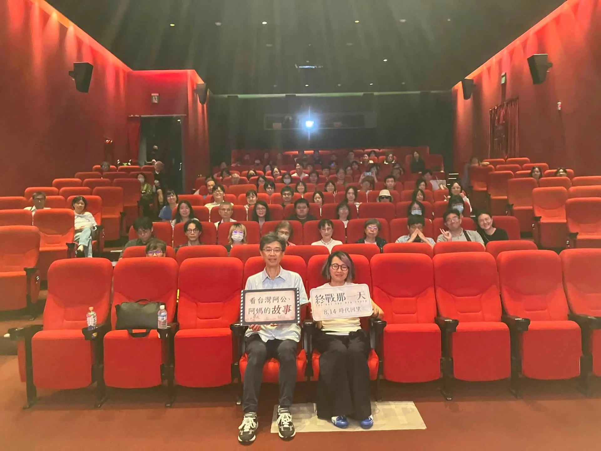 歷史顧問藍適齊與導演盧慈穎坐在電影院紅色的座椅上,手中各拿著印有電影宣傳標語的看板合影。