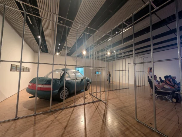 英國海沃德美術館的展場內部空間，設置有金屬鷹架結構、充氣老車裝置藝術，以及多位觀眾坐在電競椅上觀看螢幕。