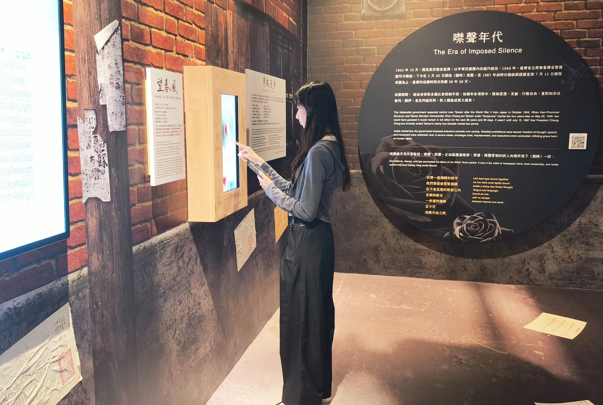 一名女性觀眾在展場互動裝置前,一邊戴著耳機聆聽語音導覽,一邊操作觸控螢幕。
