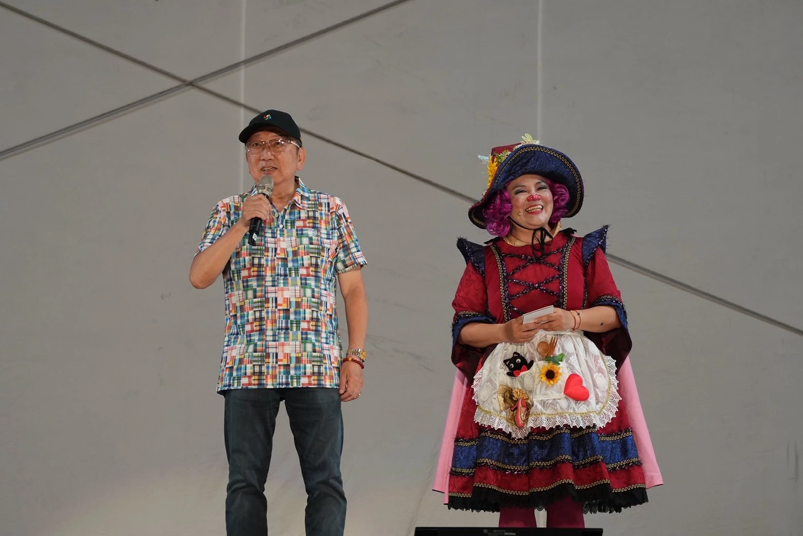一名戴黑色帽子與格子襯衫的男子在台上拿著麥克風致詞,旁邊站著一位穿著紅色華麗宮廷服飾與小丑帽的女性表演者。