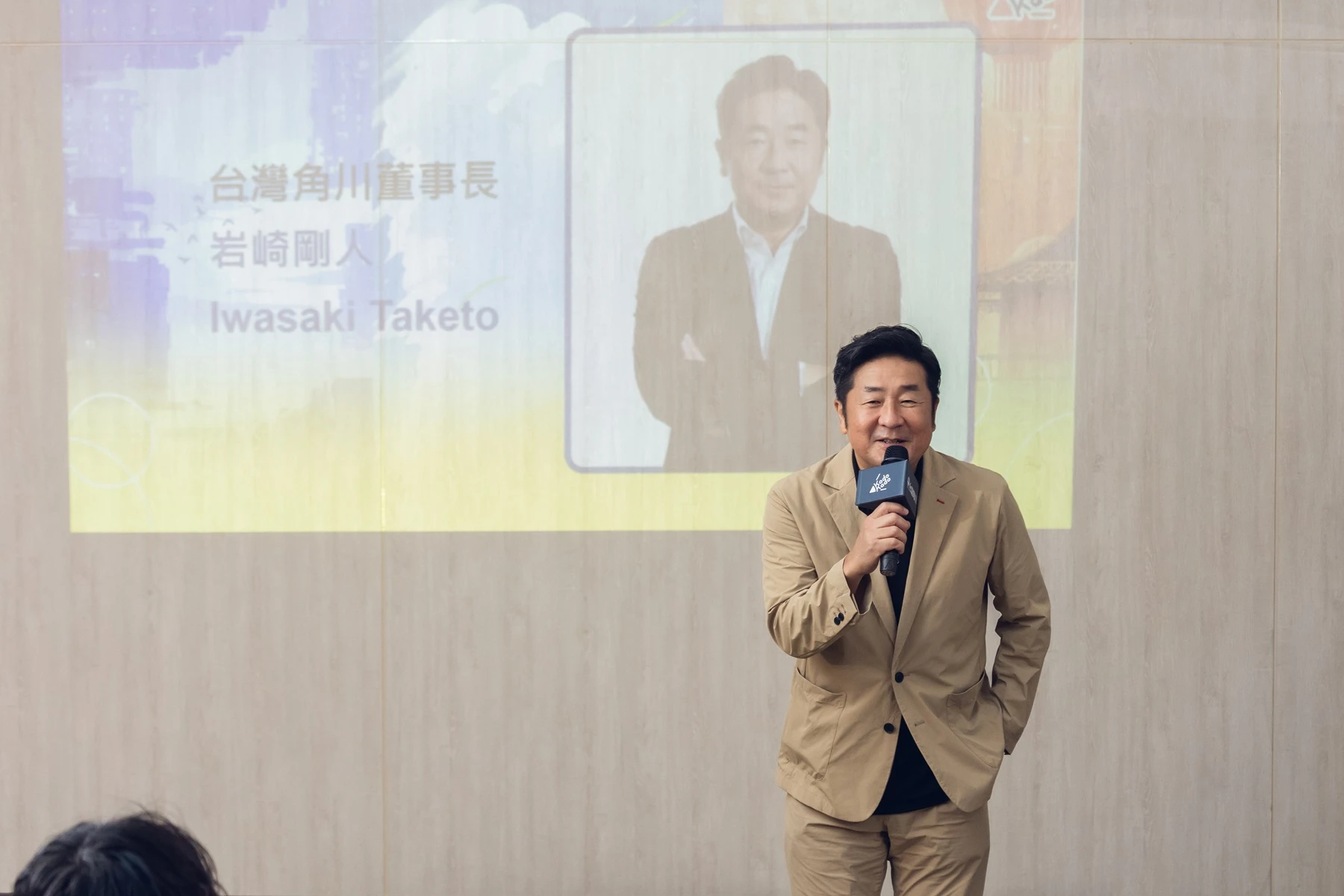 台灣角川董事長岩崎剛人在講台上持麥克風致詞,背景投影幕顯示其肖像與職稱。