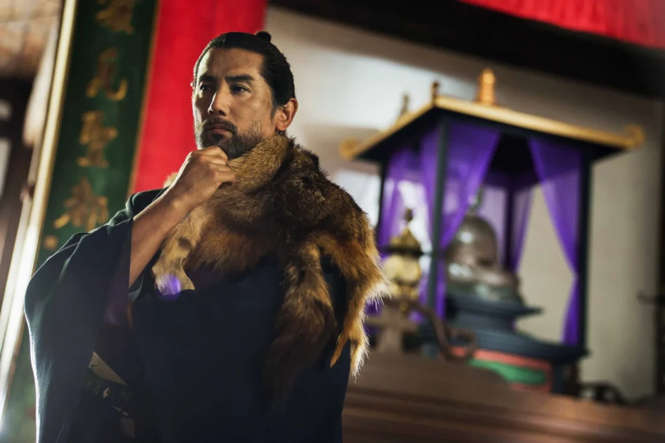 身穿深色古裝與毛皮披肩的男子，神情凝重地將手放在下巴上思考。