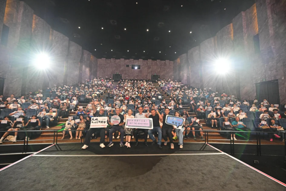 戲院放映廳內坐滿觀眾，前方舞台站有多位電影主創人員與觀眾一同面向鏡頭舉起標語合影。