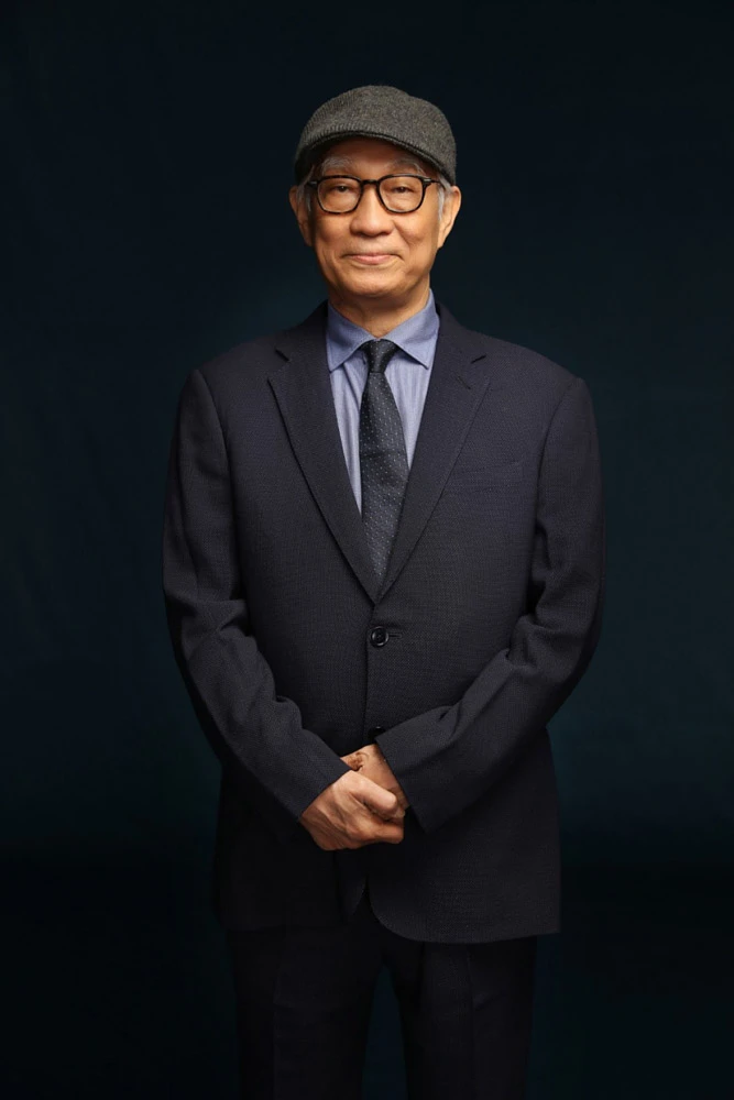 戴著鴨舌帽與眼鏡的男子廖慶松身穿深色西裝與藍色襯衫,雙手交疊站立。