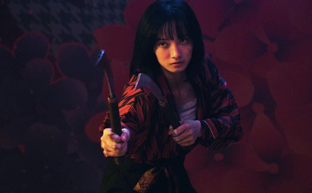 韓寧身穿紅黑印花服裝,雙手各持一把短鐮刀擺出武打姿勢,神情銳利。