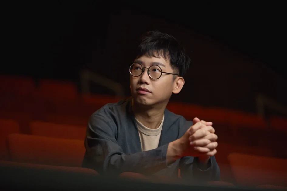 戴著圓框眼鏡的男子劉國瑞坐在紅色的劇院座椅前，雙手交疊在胸前。