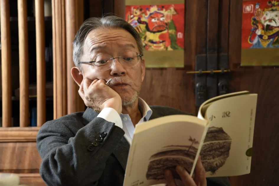 戴著眼鏡的年長男子正低頭翻閱一本書，左手托著下巴並夾著一枝筆。