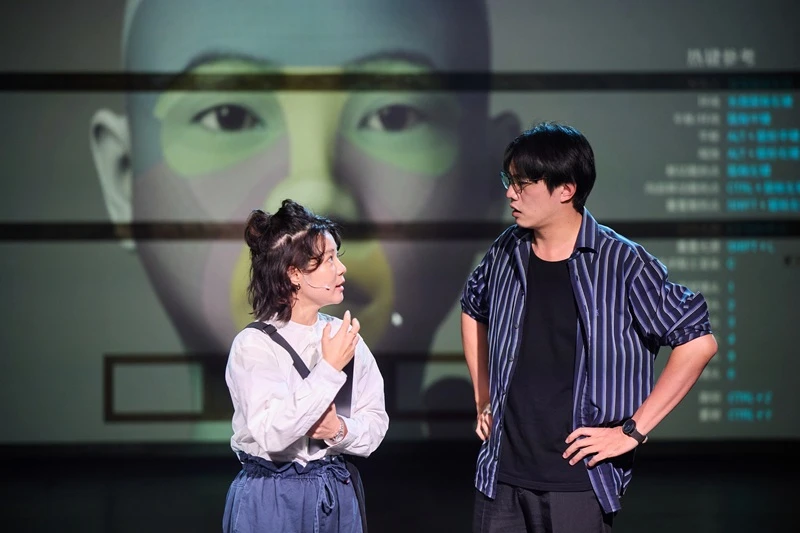 兩名演員站在劇場舞台上對話,背景大螢幕顯示人臉圖像與文字介面。