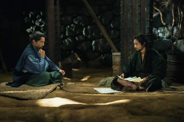 兩名古代日本武士打扮的男子坐在陰暗的地牢中，左側男子托腮沉思，右側男子盤腿閱讀手中文件。