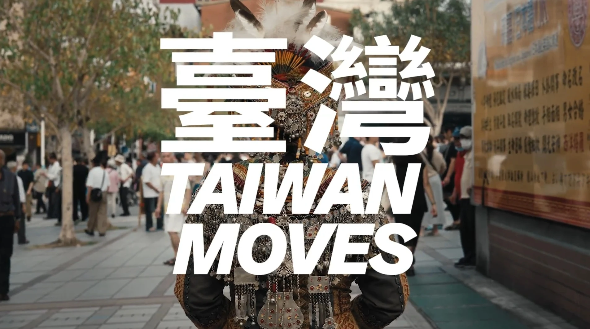 背對鏡頭的人物身著具臺灣原住民風格的華麗傳統服飾，站在熱鬧街頭上，前方疊印白色「臺灣 TAIWAN MOVES」字樣。