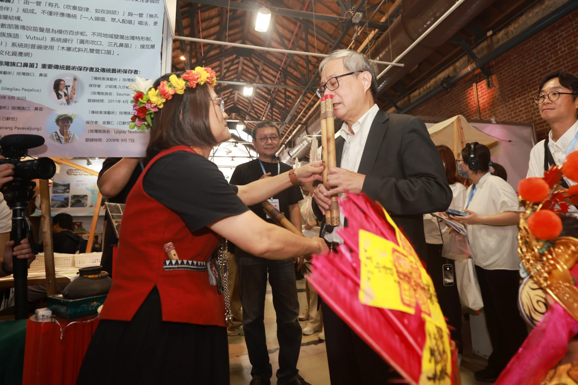 文化部長李遠拿著竹製鼻笛體驗吹奏,前方有穿著原住民傳統服飾的女性協助引導。