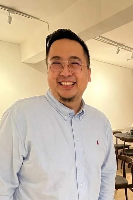 穿著淺藍色長袖襯衫與細框眼鏡的男子面對鏡頭微笑