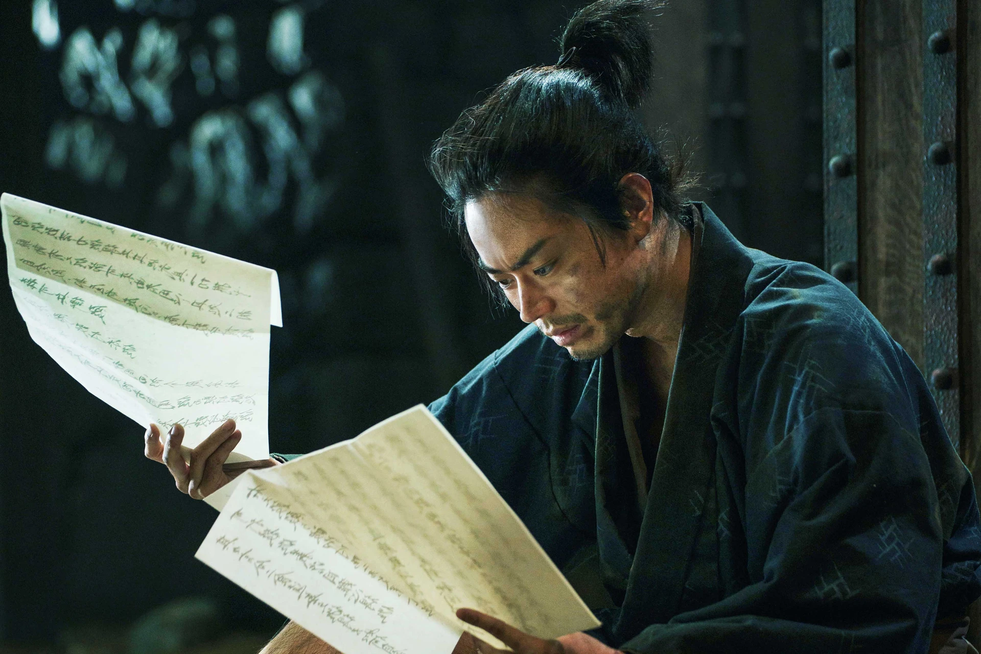 一名古代男子在昏暗光線下,神情專注地低頭閱讀手中展開的古老書信。